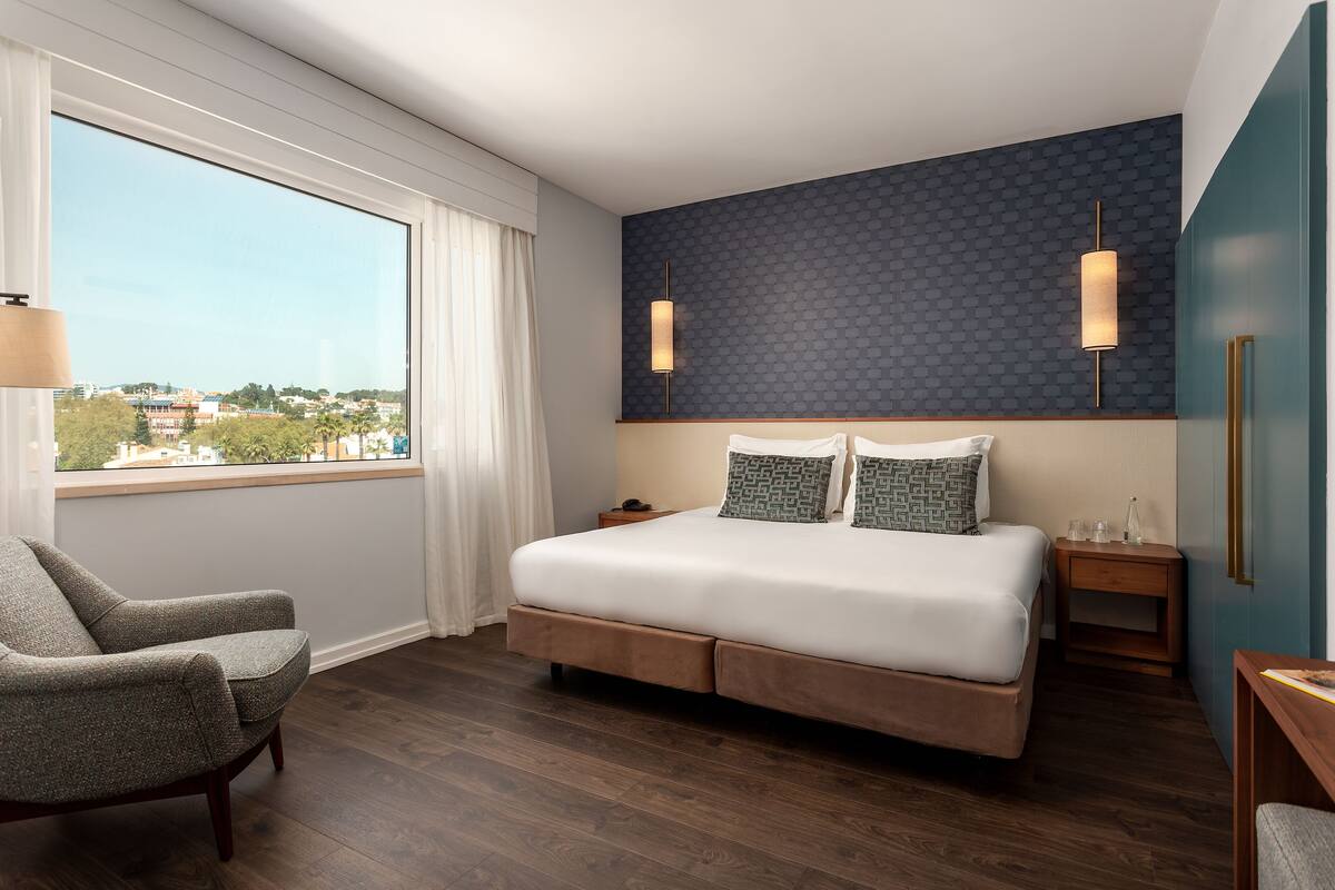 A modern hotel room with a large bed, a chair, a nightstand, and a view of the city through a large window.