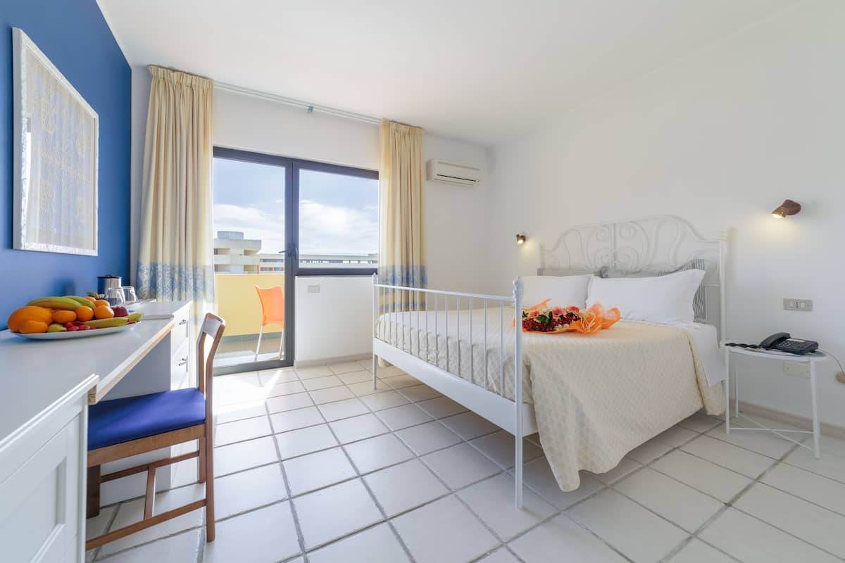 A hotel room with a bed, a desk with a chair, a small table, and a balcony with curtains.