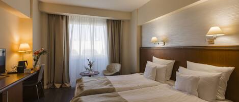 A hotel room with two beds, a desk with a lamp, a chair, and a small table with a vase of flowers.