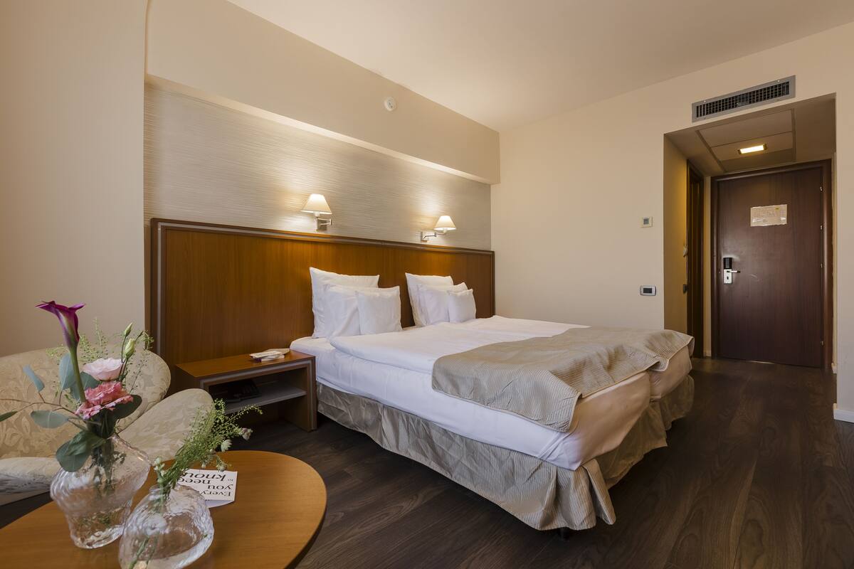 Quarto de hotel com uma cama grande, luminárias de cabeceira, um painel de madeira na cabeceira, uma mesinha de cabeceira, um vaso com flores e uma placa na mesinha.