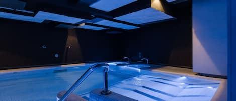 A modern indoor swimming pool with clear water, a metal handrail, and a tiled floor.