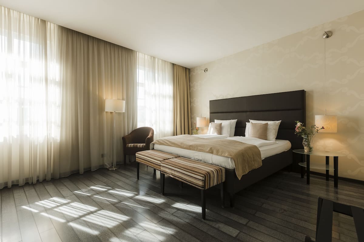 Quarto de hotel moderno com uma cama grande, um banco, duas mesas de cabeceira com luminárias, uma poltrona e cortinas translúcidas que permitem a entrada de luz natural.