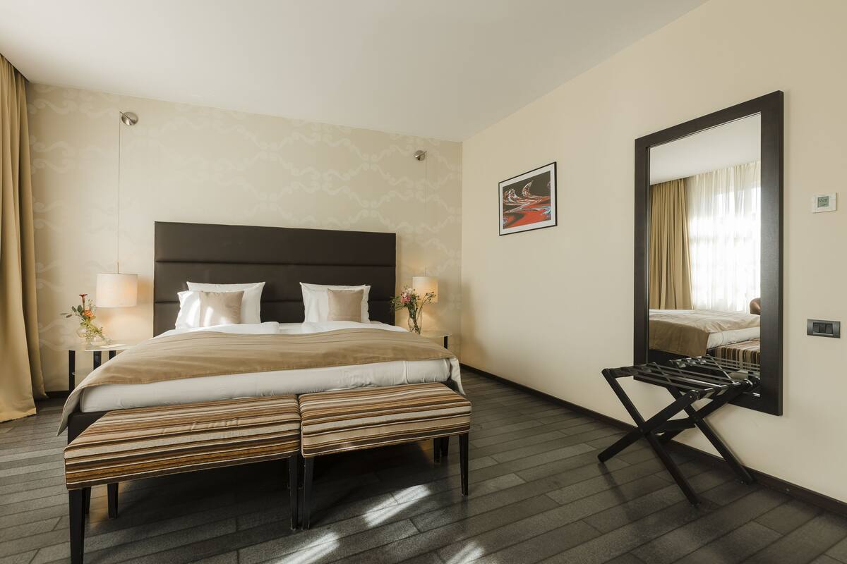 Quarto de hotel com uma cama grande, luminárias laterais, um espelho e um banco.