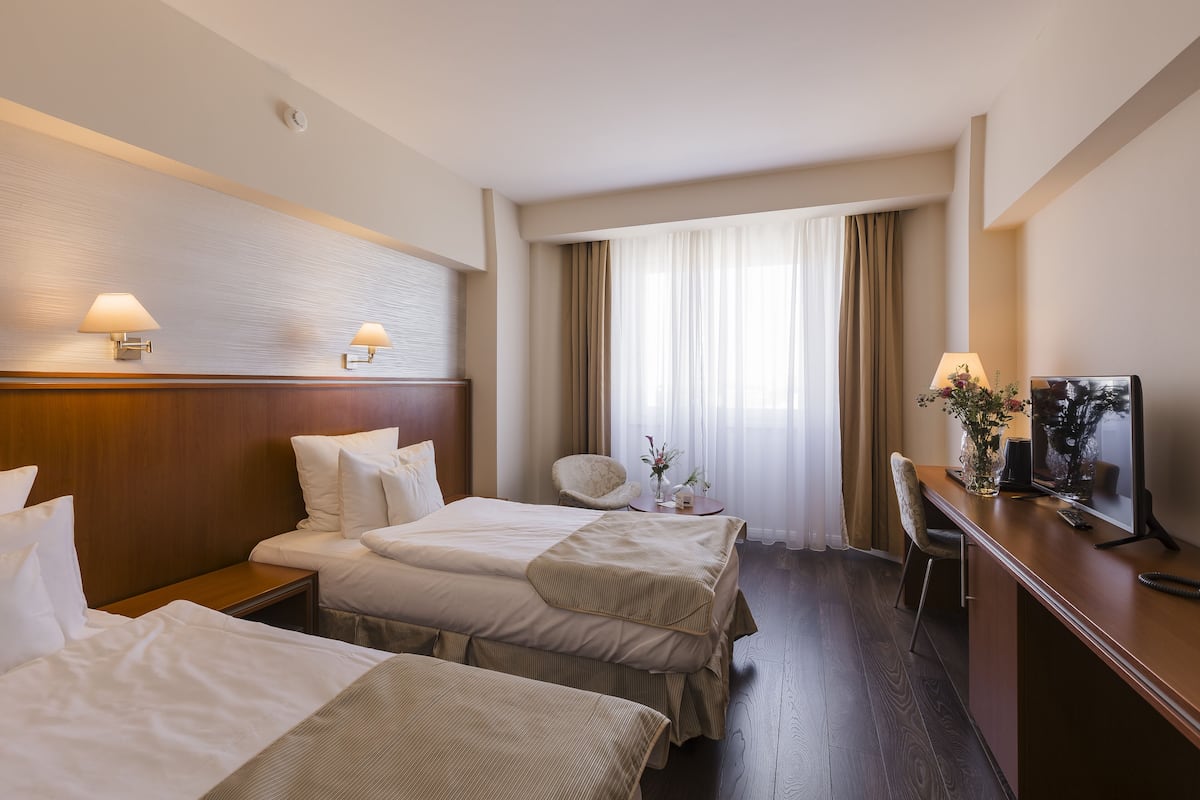 Quarto de hotel com duas camas, uma mesa, uma cadeira, televisão e janela com cortinas.