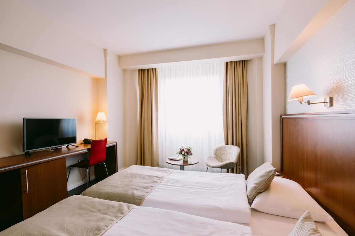 Quarto de hotel com duas camas, uma mesa de escritório com televisão, uma cadeira, uma pequena mesa com um vaso e cortinas.