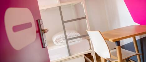 A bunk bed with a wooden desk and chair, a white drawer, and a pink wall.