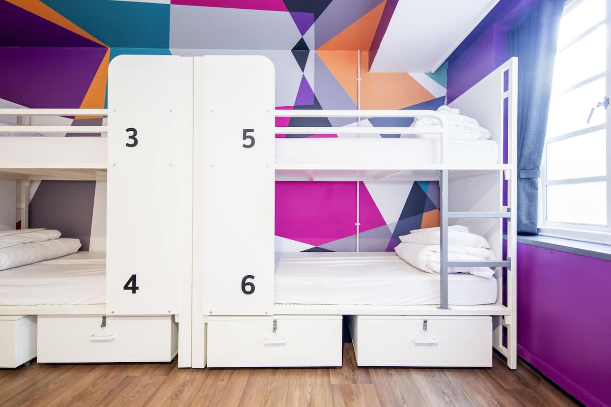 A room with bunk beds numbered 3, 4, 5, and 6, wooden flooring, and a window with curtains.