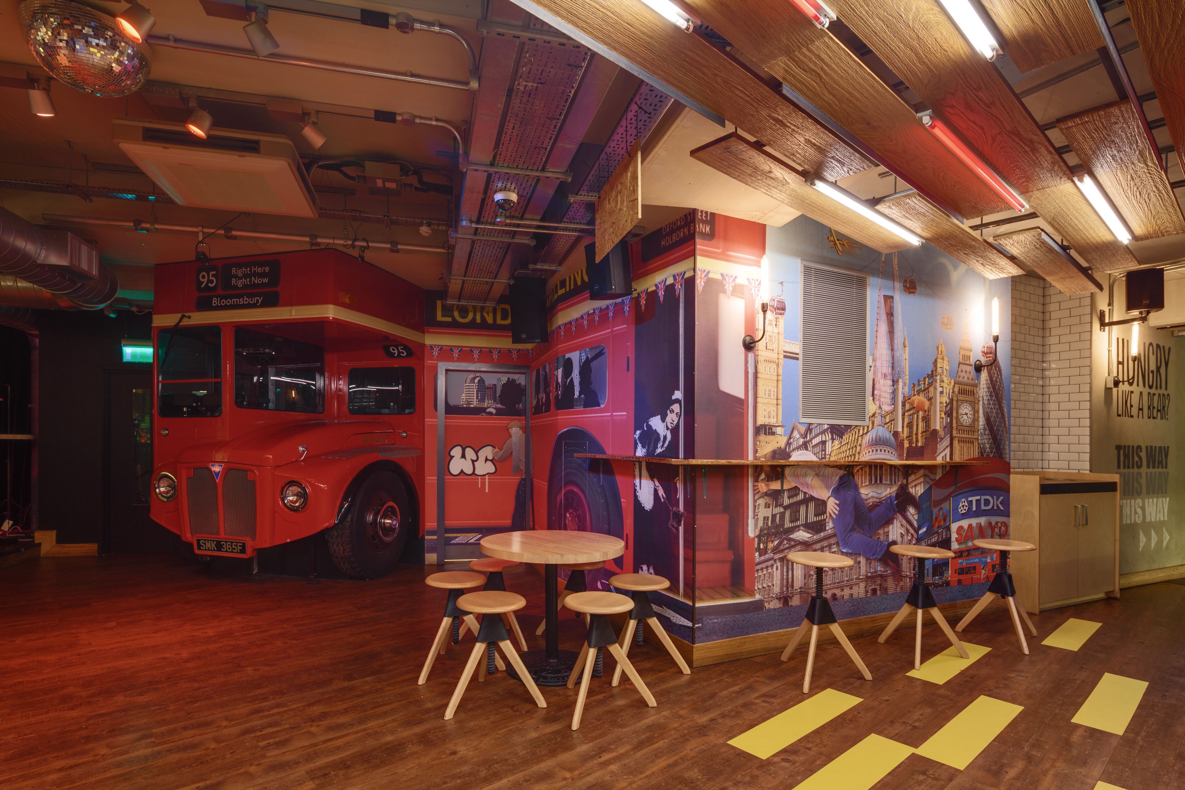 A restaurant with a red double-decker bus theme, wooden tables, and a brick wall mural.
