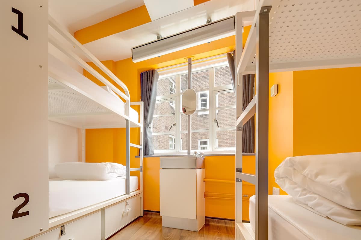 A dormitory room with bunk beds, a window with curtains, a radiator, and a kitchenette.