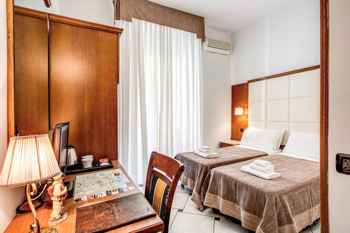 Ein Hotelzimmer mit zwei Betten, einem Schreibtisch mit Lampe, einer Karte und einem Holzschrank.