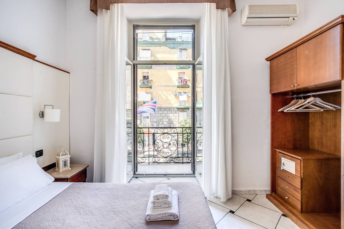 Ein Hotelzimmer mit Bett, einem Holzschrank, einem Balkon mit Blick auf Gebäude und einer Klimaanlage.
