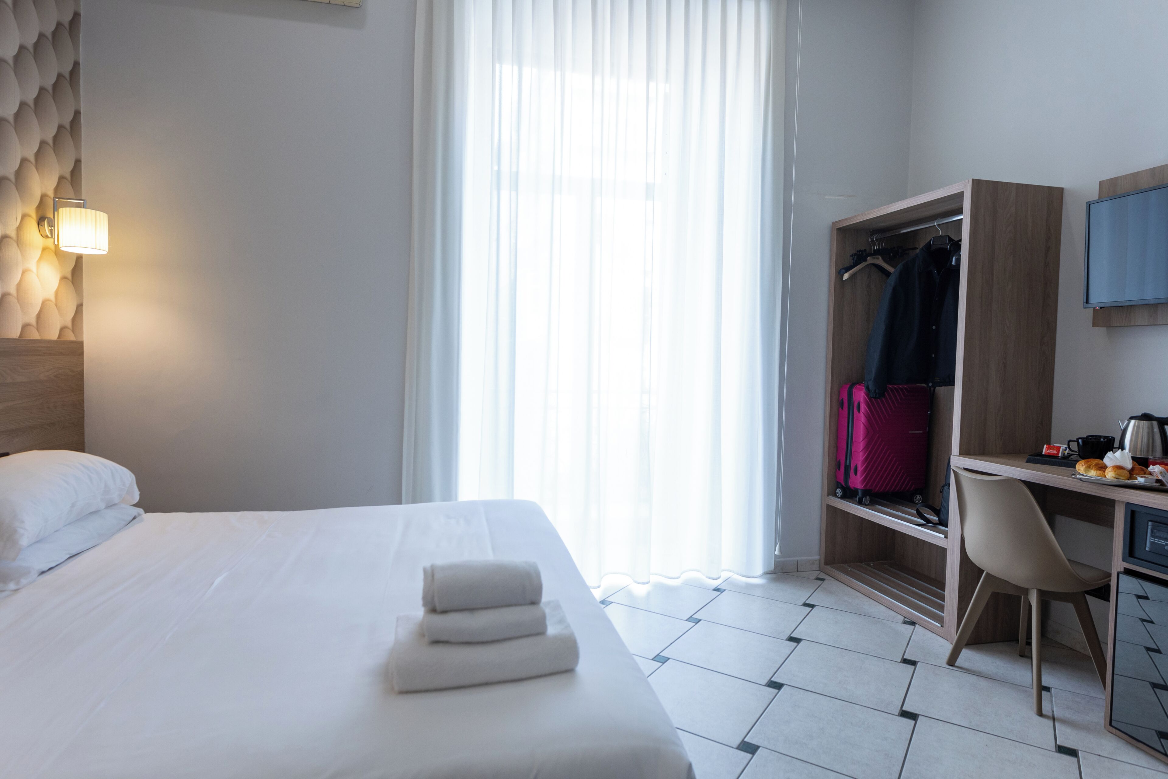 Habitación de hotel con cama, escritorio, silla, armario y ventana con cortinas.