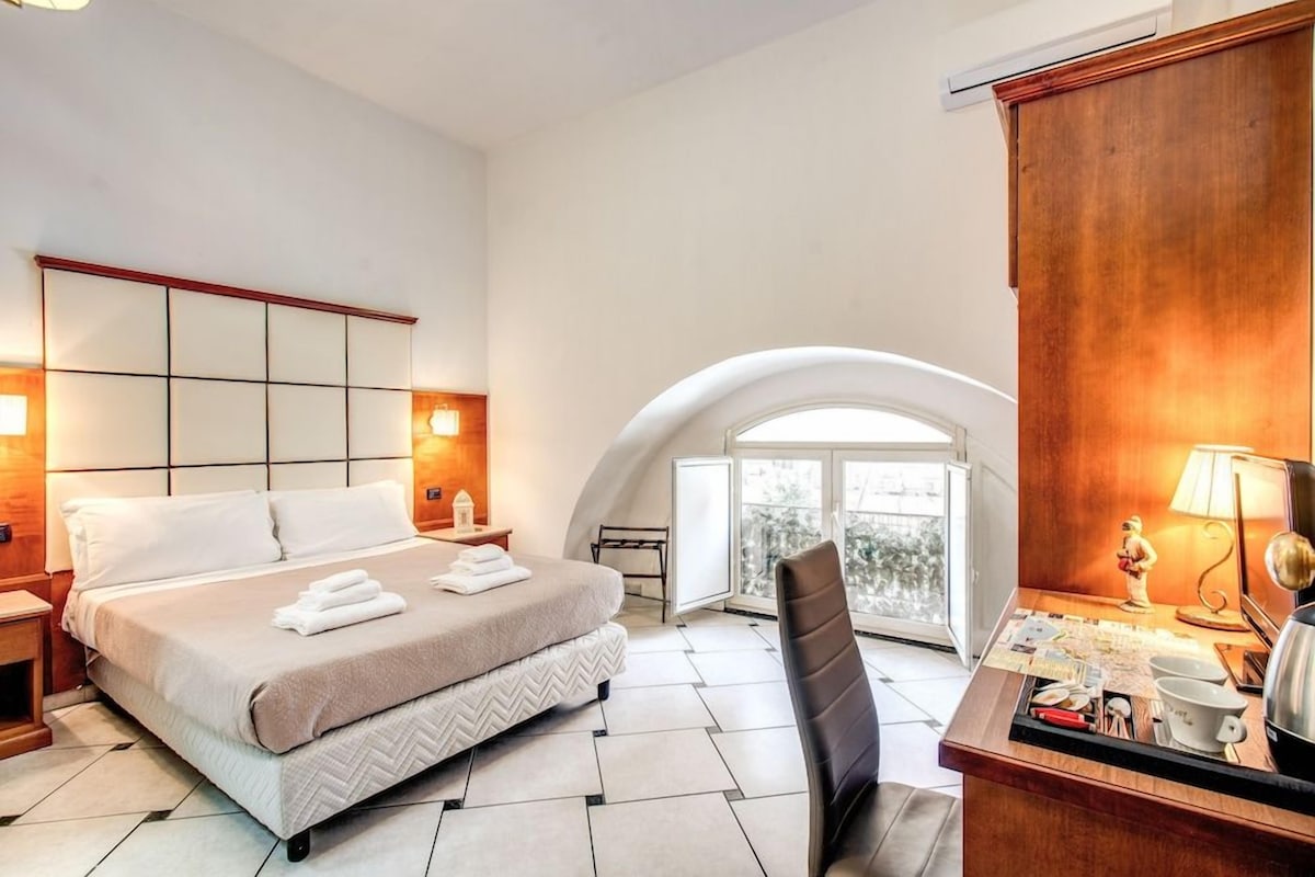 A hotel room with a bed, a desk with a lamp, a chair, and a view through an arched window.