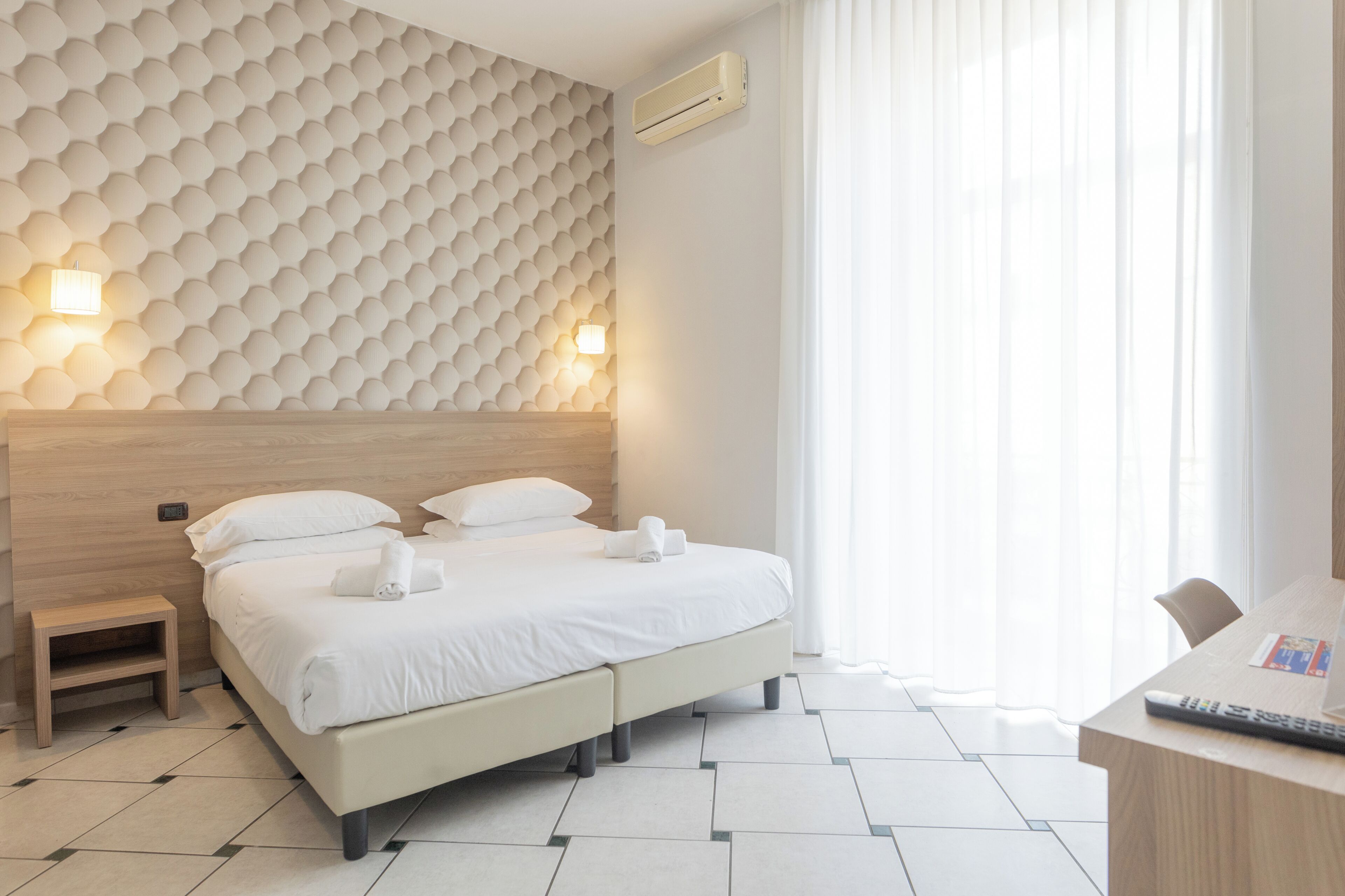 A hotel room with a bed, bedside tables, a desk, and a window with curtains.
