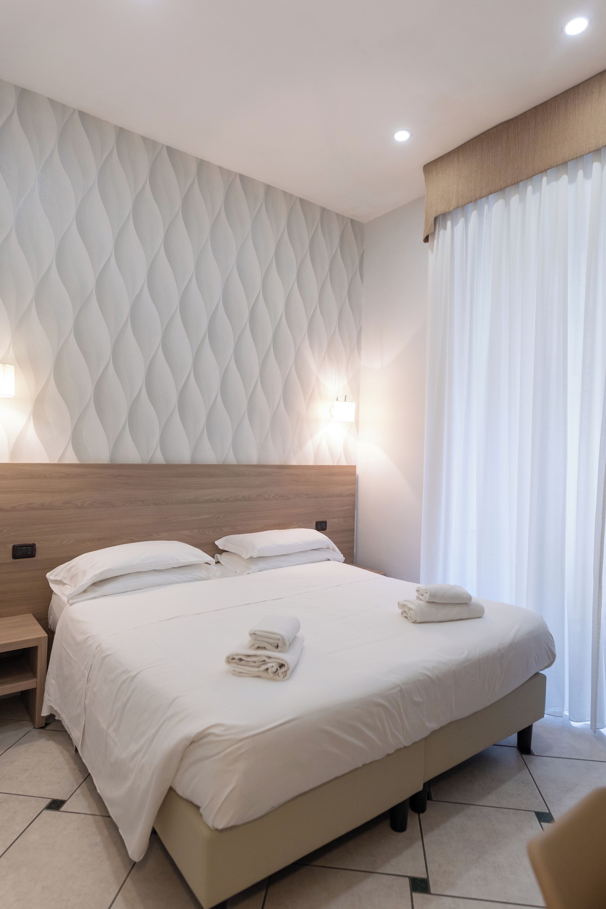 Habitación de hotel con una cama grande, dos mesitas de noche, un cabecero y una ventana con cortinas.