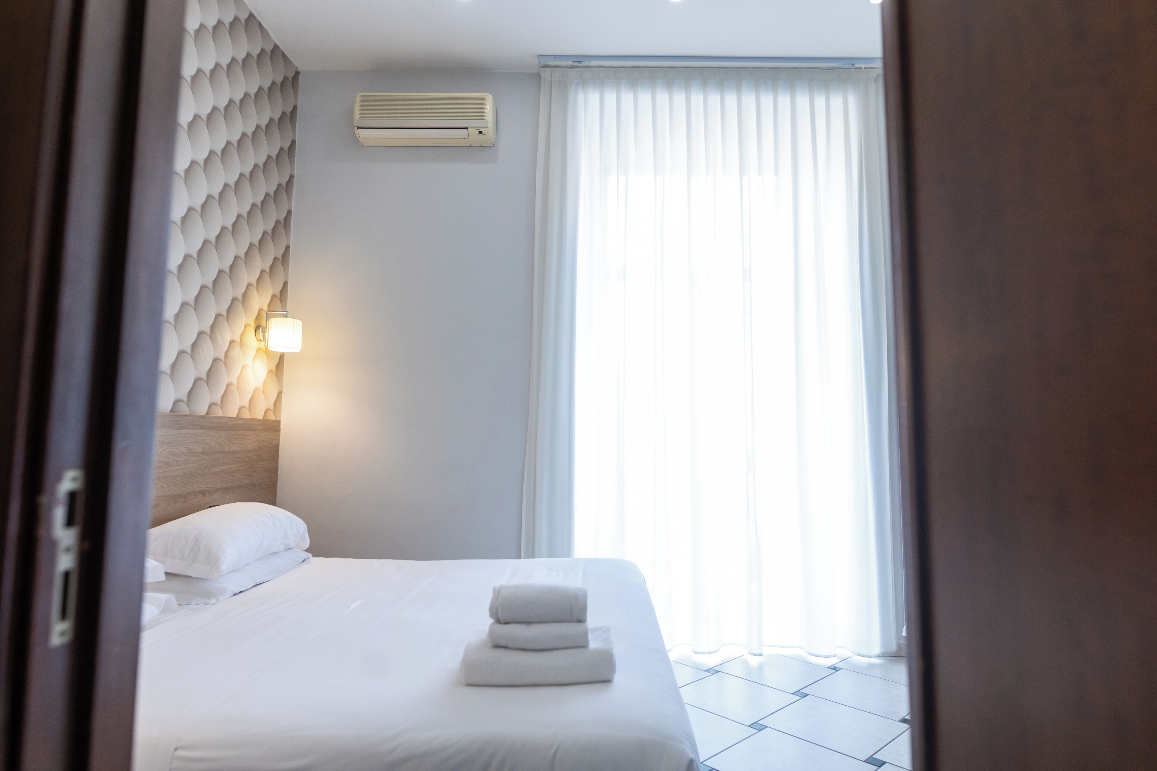 A hotel room with a bed, a wall-mounted air conditioner, a bedside lamp, and a window with curtains.