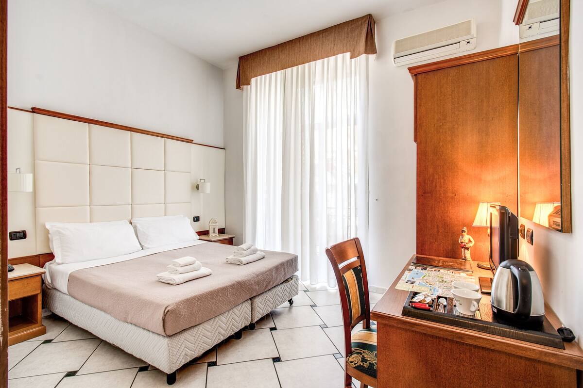 Ein Hotelzimmer mit einem großen Bett, einem Holztisch, einem Stuhl, einer Schreibtischlampe, einem Wasserkocher und einer Karte auf dem Tisch.