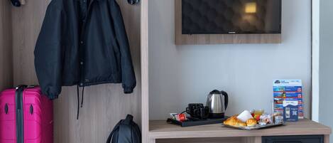 A hotel room with a wooden wardrobe, a desk with a coffee maker, a chair, a pink suitcase, a black backpack, and a television on the wall.
