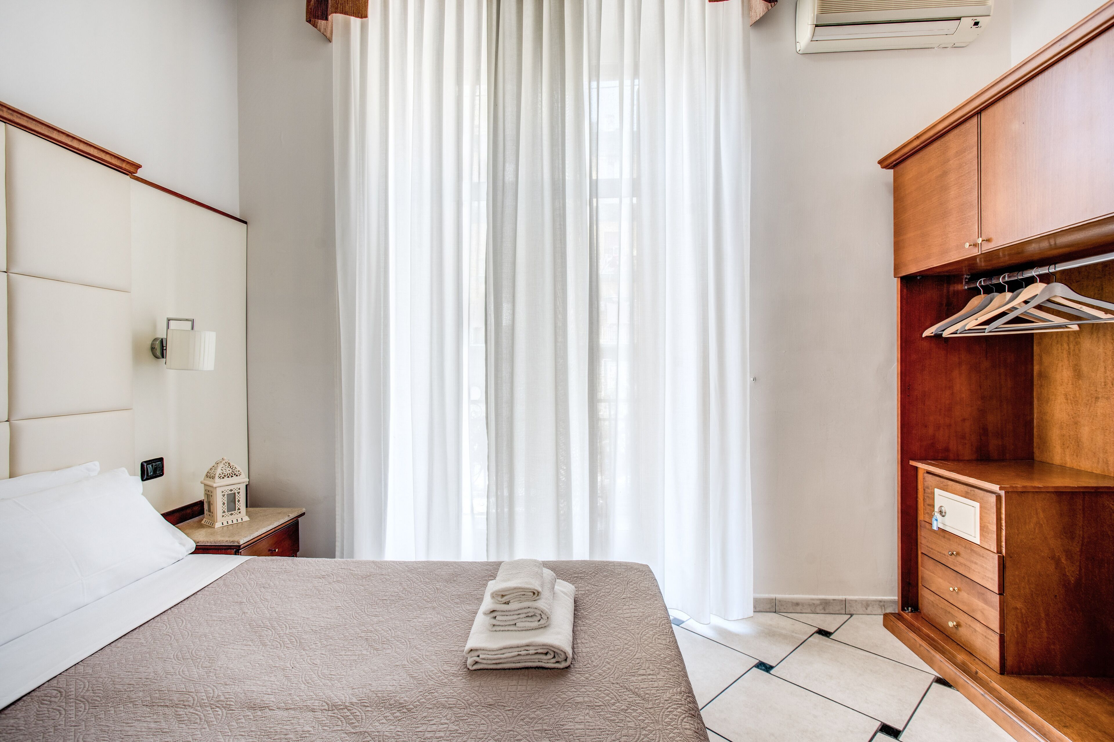 A neatly made bed with a folded towel, a wooden wardrobe with hangers, and a nightstand with a lamp and a decorative item.