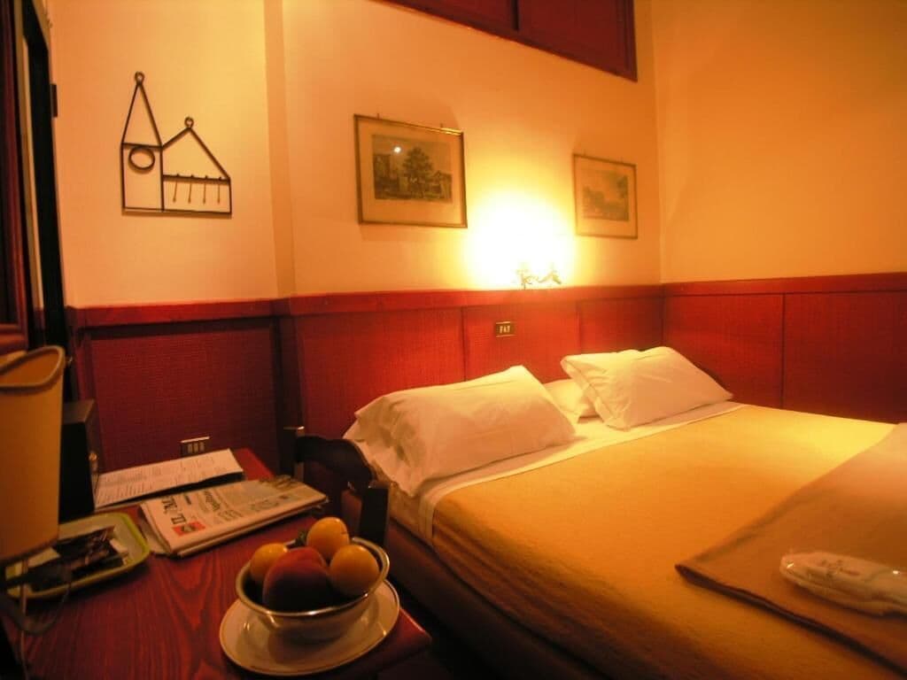 A hotel room with a bed, a nightstand with a lamp, a bowl of fruit, and a wall with a picture and a sign.