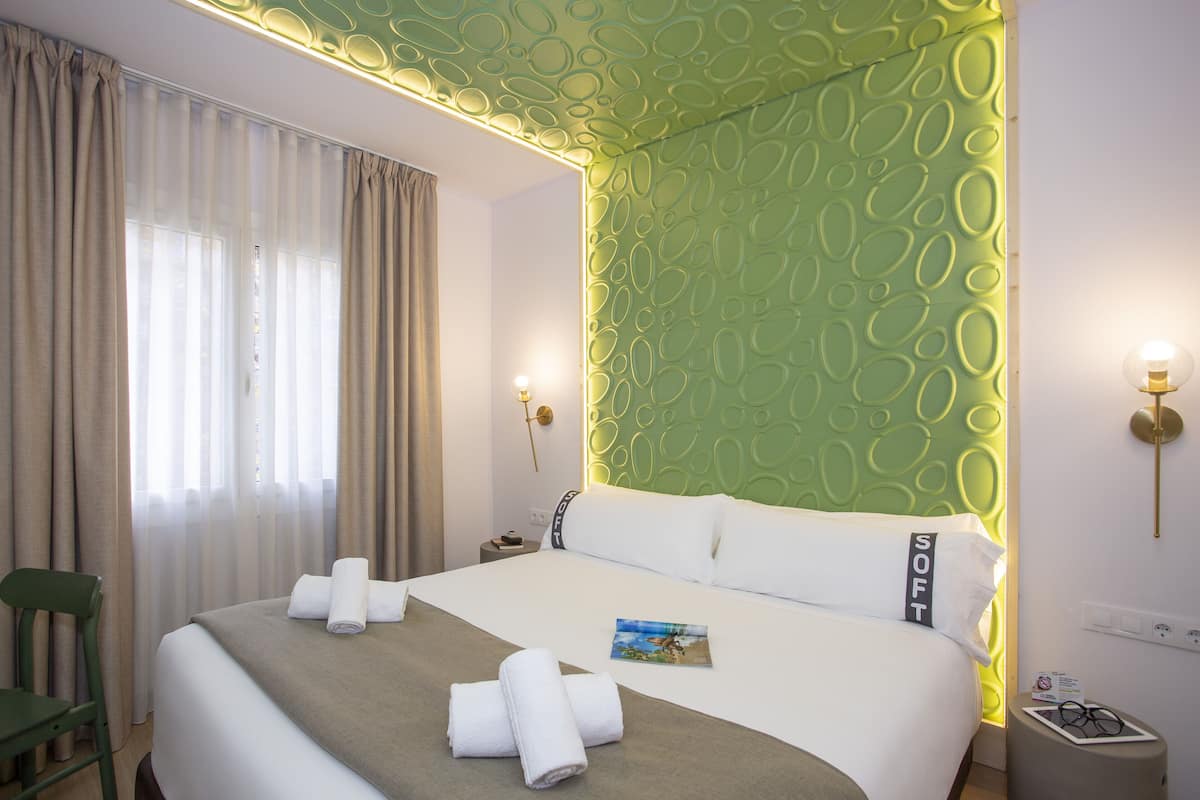 Habitación de hotel con cama, cortinas, una silla, una mesita de noche con lámpara y una pared verde con un diseño circular.