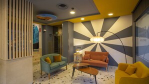 A modern living room with a terrazzo floor, a blue sofa, an orange sofa, and a yellow armchair. There is a floor lamp and a small table with a lamp. - Casual Colours Barcelona Hotel by Casual Hoteles (Barcelona)