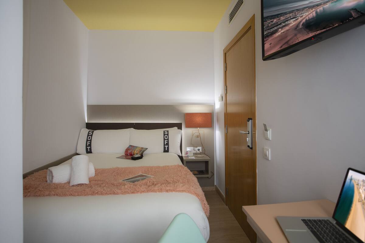 Una habitación de hotel con una cama, un escritorio con una computadora portátil, un televisor en la pared y una mesita de noche con una lámpara.