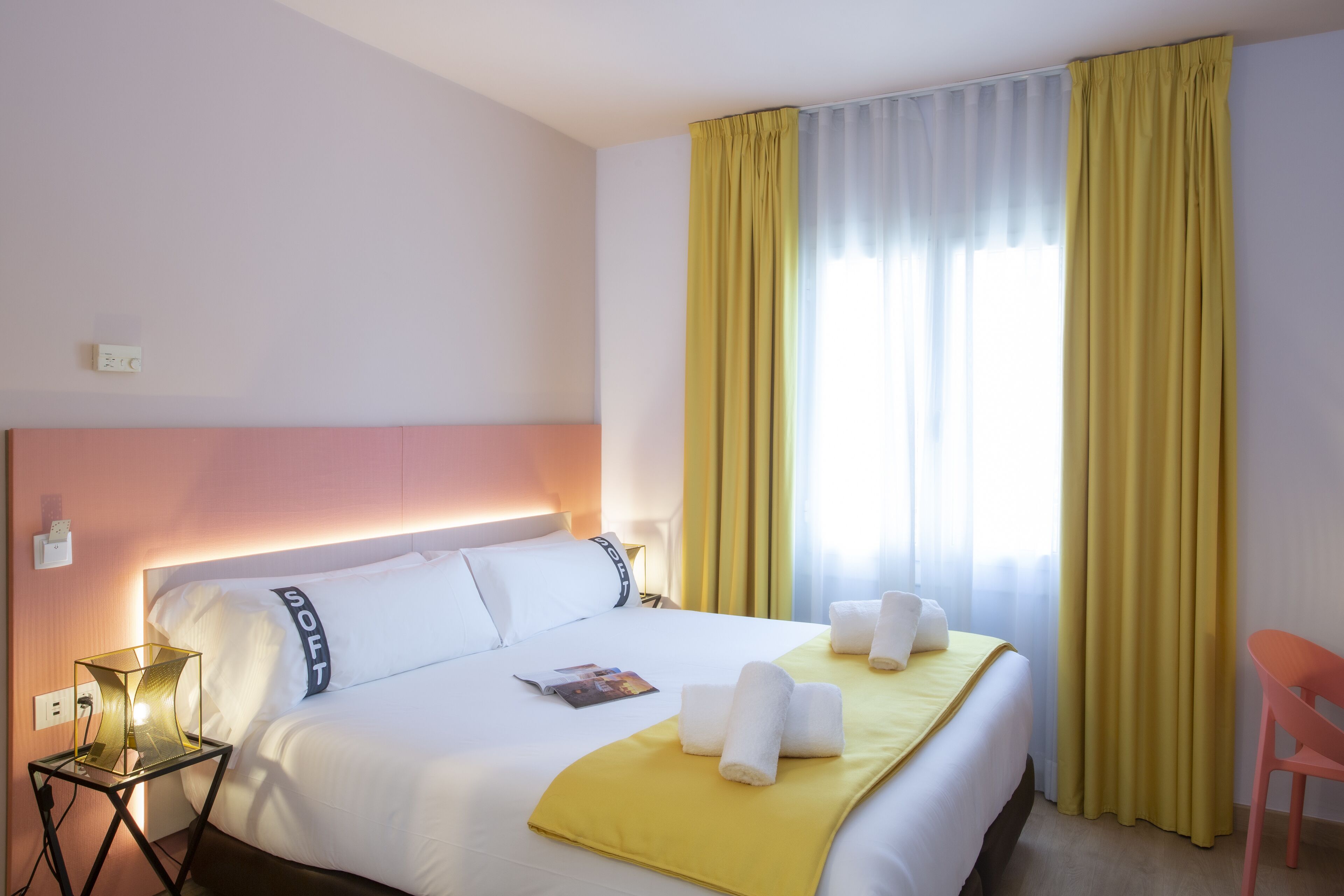 Quarto de hotel com cama, mesa de cabeceira com abajur, cadeira e janela com cortinas.