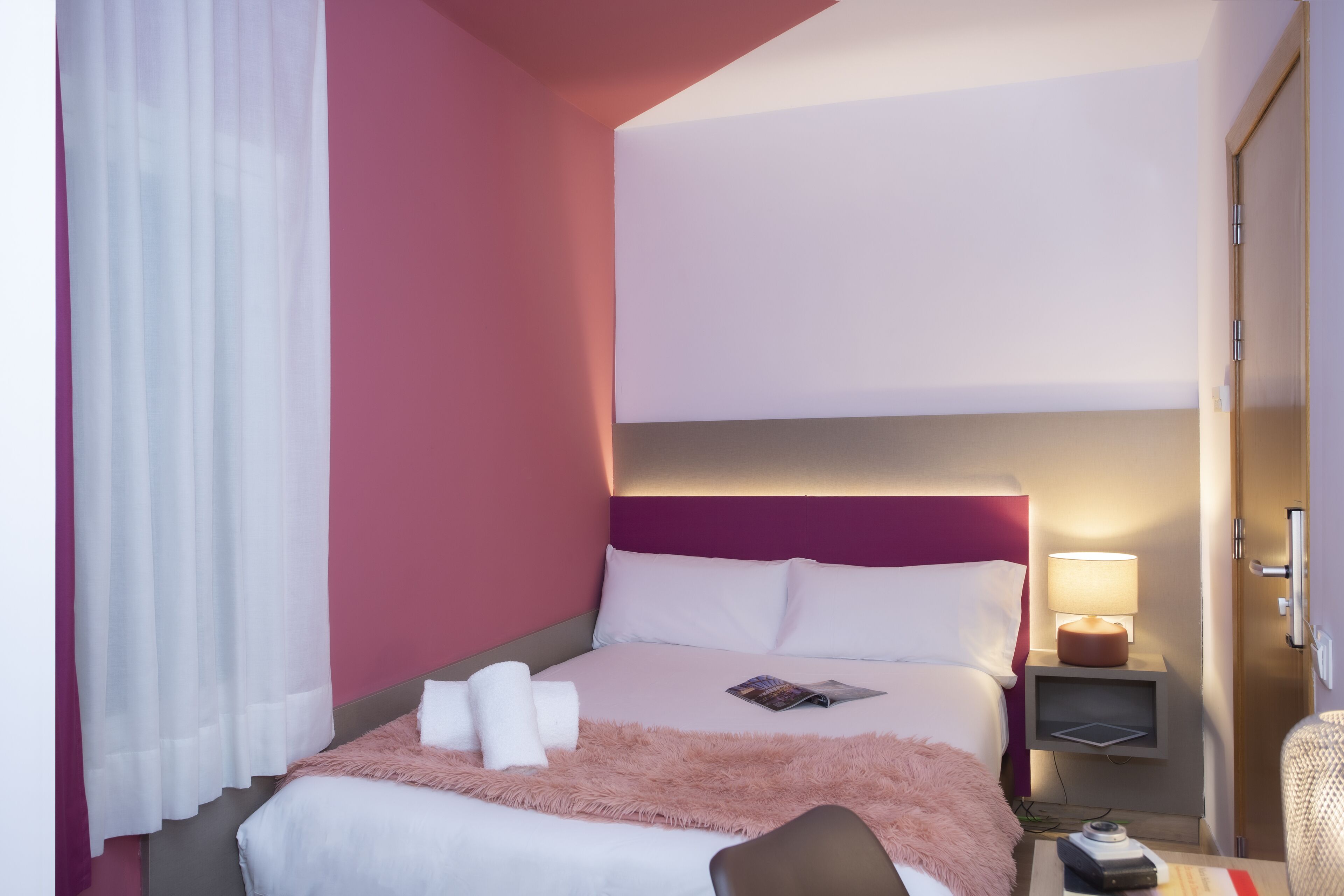 A hotel room with a bed, a nightstand with a lamp, and a door.