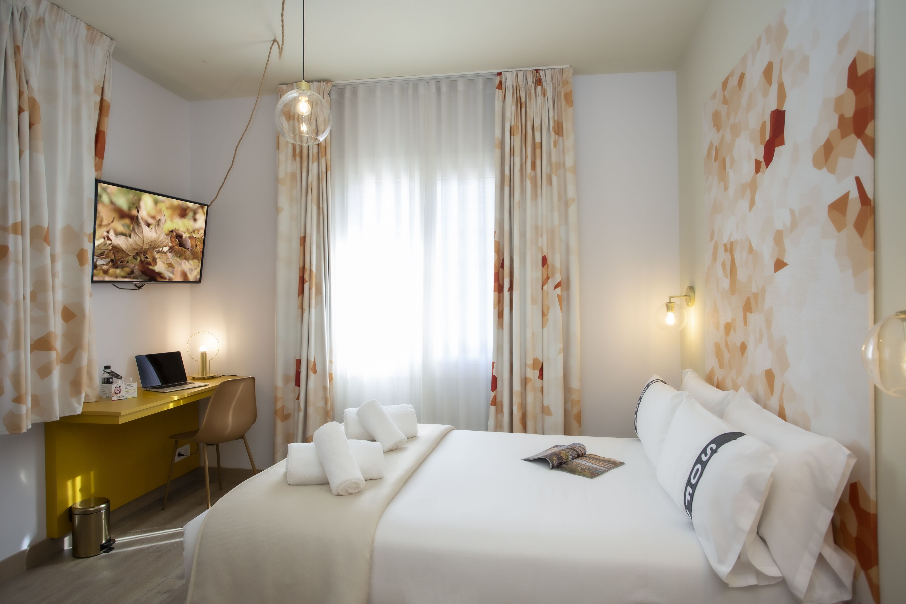 A hotel room with a bed, a desk with a laptop, a chair, and a TV mounted on the wall.