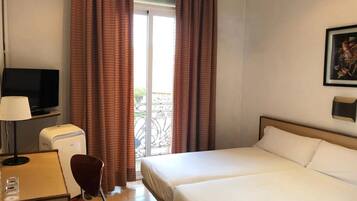 Habitación de hotel con cama, escritorio con lámpara, televisión y balcón con cortinas.