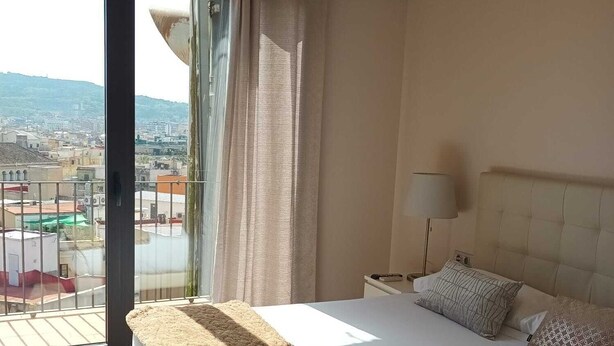 Een hotelkamer met een groot bed, een nachtkastje, een lamp en uitzicht op een stadsgezicht door een groot raam.
