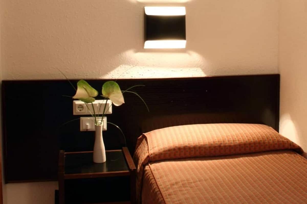 A hotel room with a bed, a nightstand, a vase with a plant, and a wall-mounted light fixture.