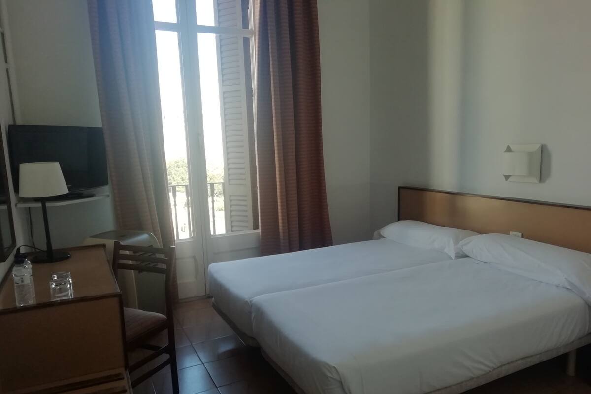 A hotel room with a bed, a desk with a lamp, a chair, a TV, and a window with curtains.