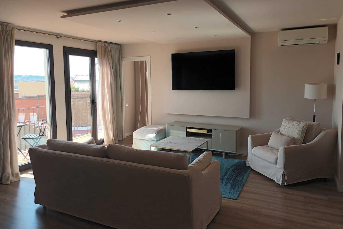 A modern living room with a sofa, armchair, flat-screen TV, and a balcony with curtains.