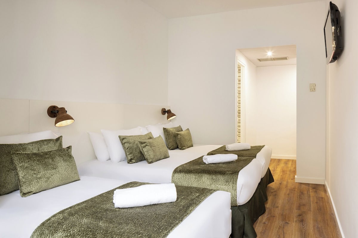 Een hotelkamer met twee bedden, elk met groene beddengoed en witte kussens. Aan beide zijden van de bedden hangen wandlampen.