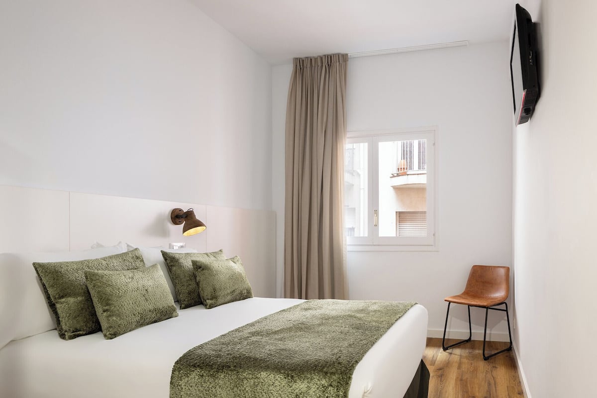 Een hotelkamer met een bed, een stoel, een raam met gordijnen en een aan de muur bevestigde televisie.