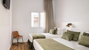 A hotel room with a bed, a chair, a TV, a window with curtains, and a wall-mounted light fixture.