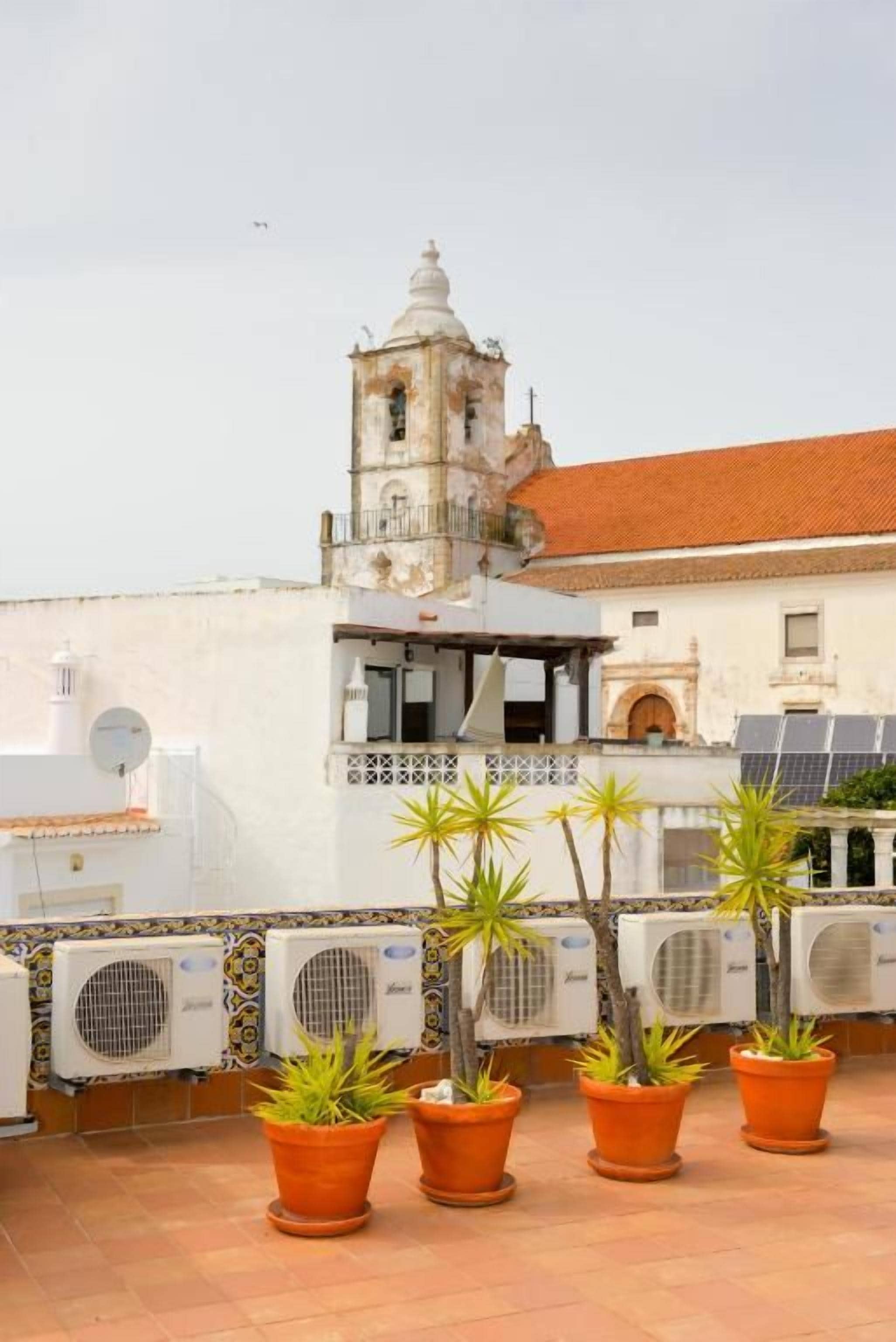 A rooftop terrace with potted plants, a bell tower, and air conditioning units.