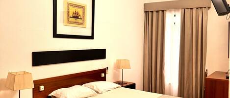A hotel room with a bed, two bedside tables, a nightstand, a lamp, a framed picture on the wall, and a window with curtains.