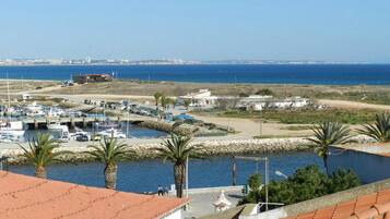 Uma marina costeira com barcos, vista clara do mar e um horizonte urbano distante.