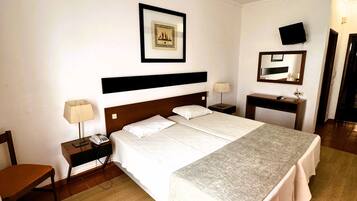 Quarto de hotel com uma cama grande, mesa de cabeceira com abajur, uma cadeira, escrivaninha com computador, televisão e um quadro na parede.