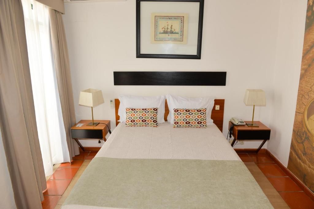 A hotel room with a bed, two bedside tables, a nightstand, a framed picture on the wall, and a window with curtains.