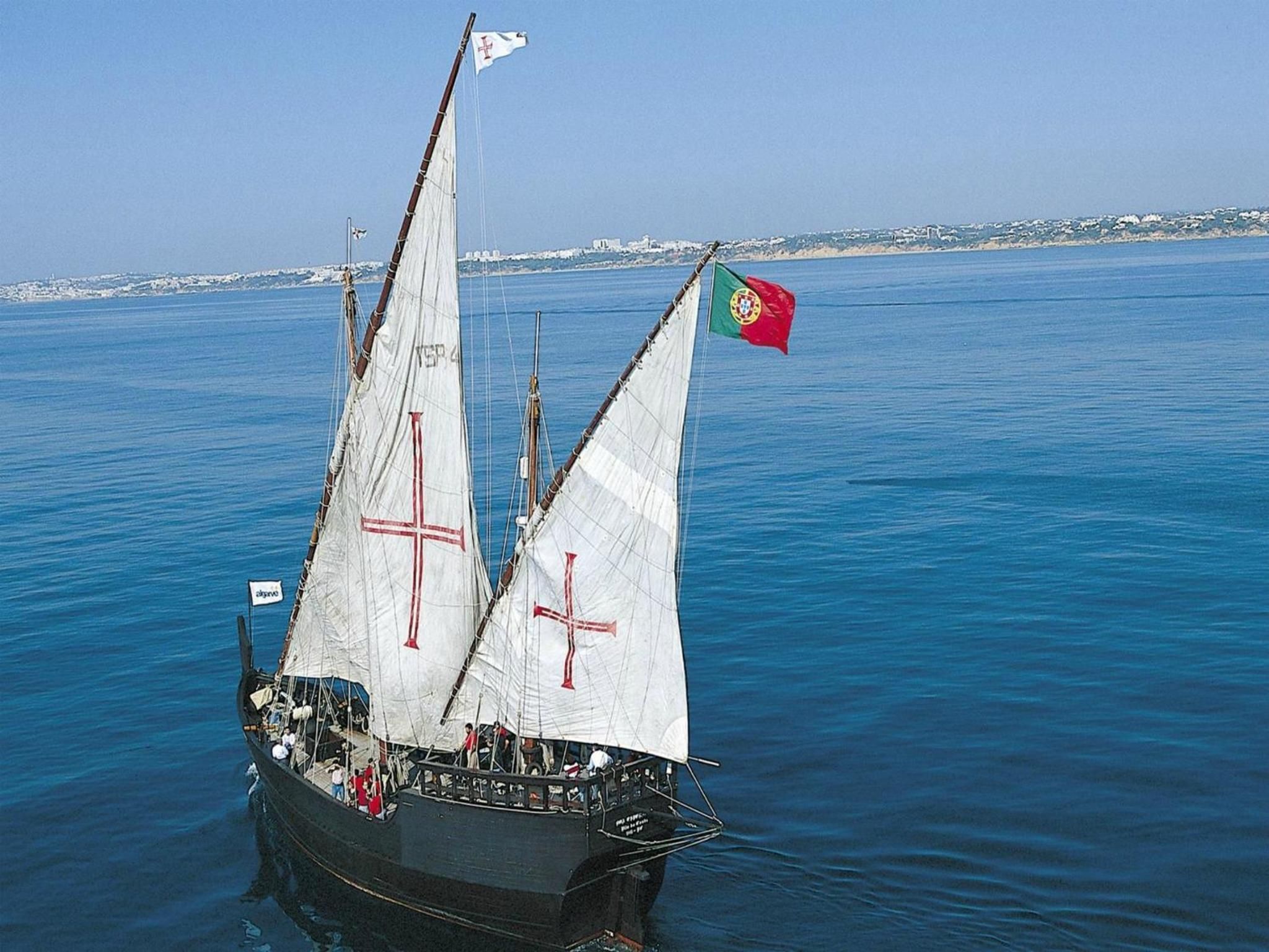 Ein Segelboot mit mehreren Segeln, einer Flagge und einer Küstenlinie im Hintergrund.