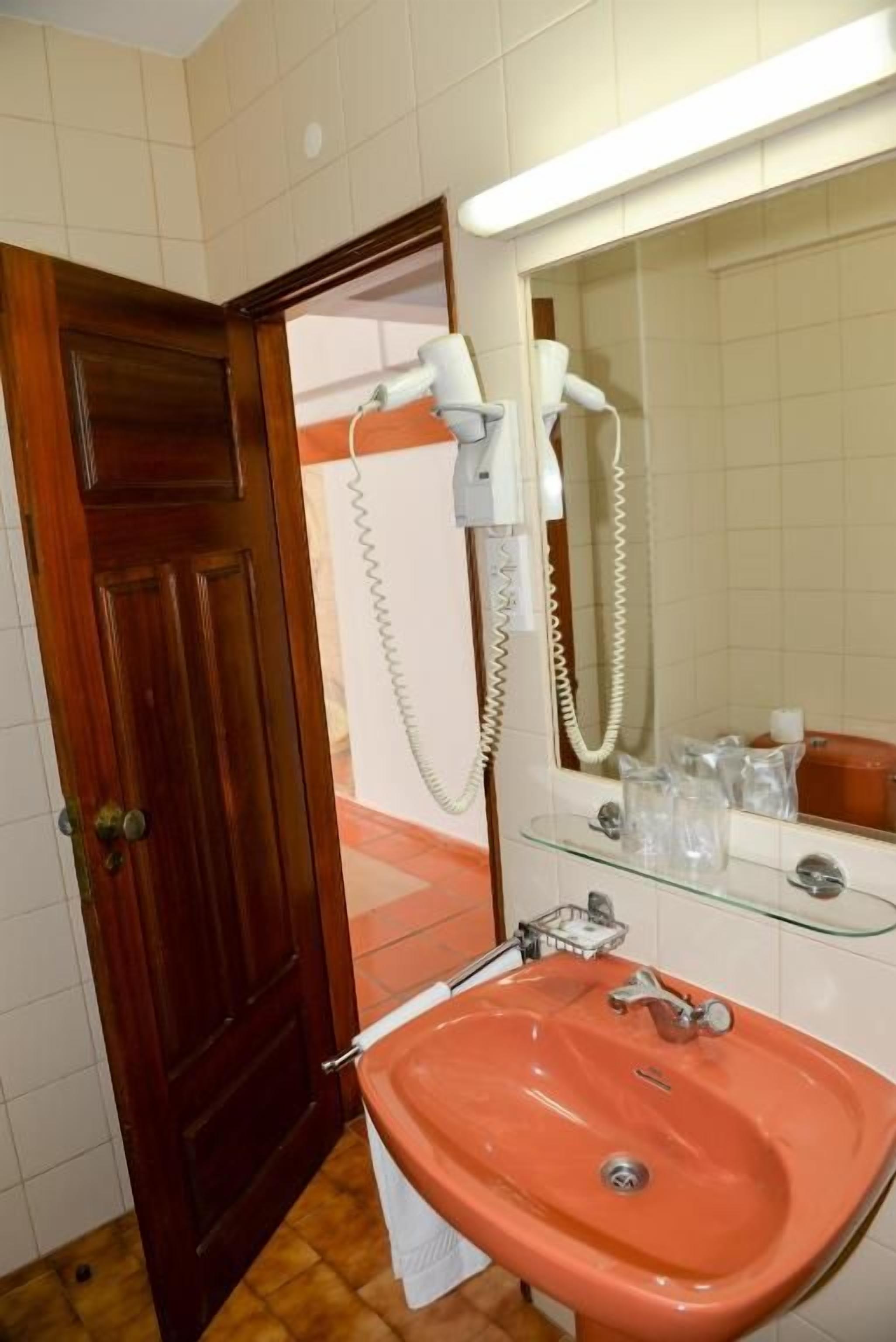 A bathroom with a wooden door, a pink sink, a glass shelf with toiletries, and a hairdryer.
