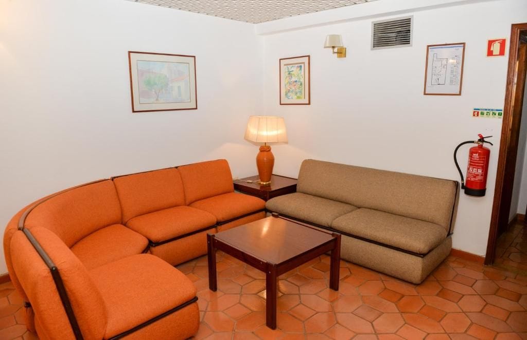 Quarto com dois sofás, uma mesinha e uma luminária. Quadros emoldurados nas paredes e extintor de incêndio fixado na parede.