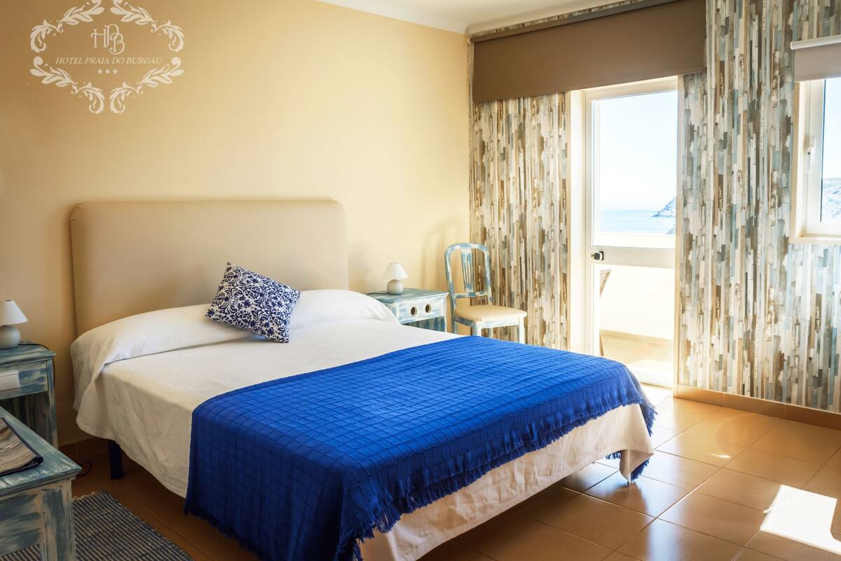 A hotel room with a bed, a nightstand, a chair, and a view of the sea through a door.