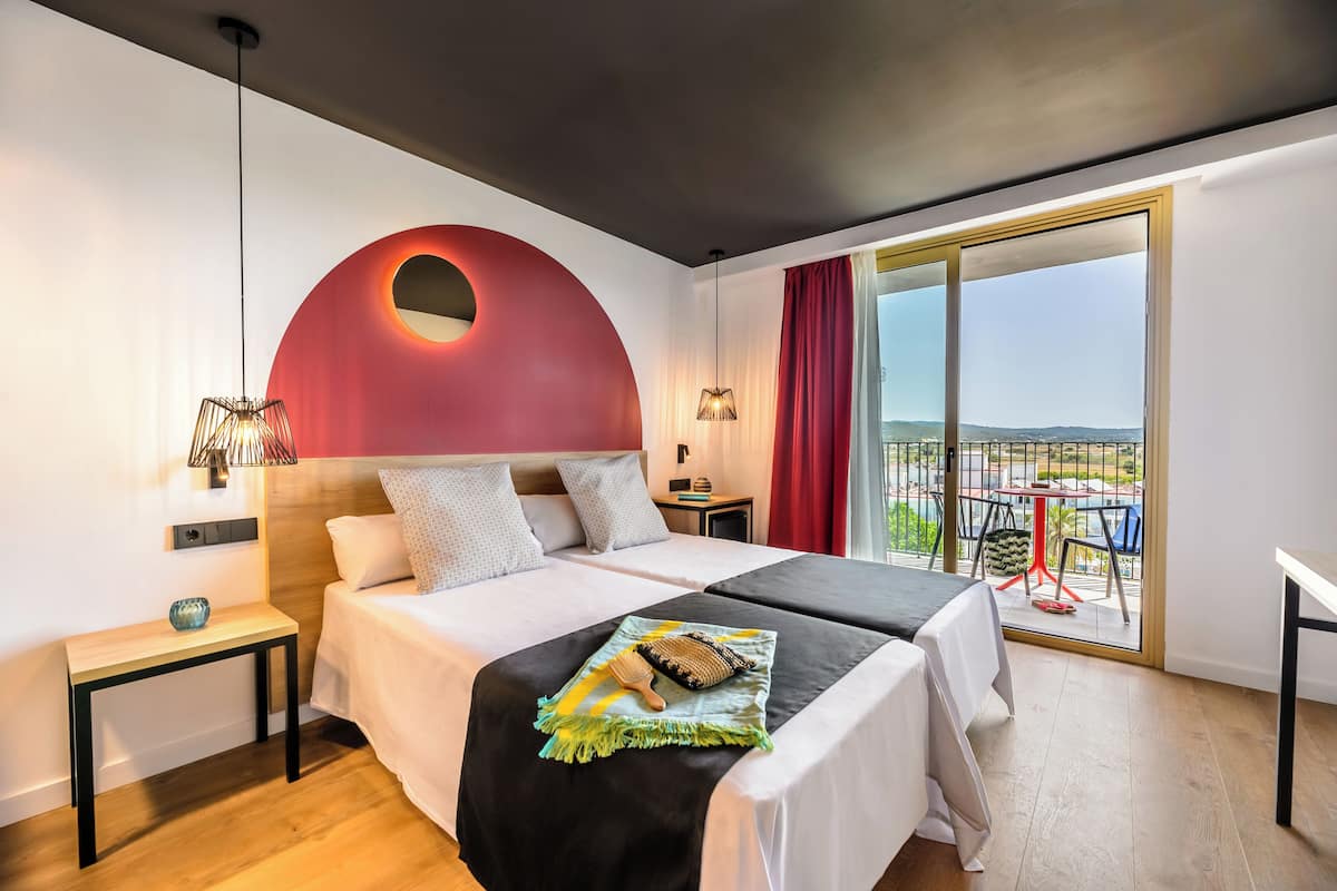 A modern hotel room with two beds, a balcony with a table and chairs, and a large window offering a view.