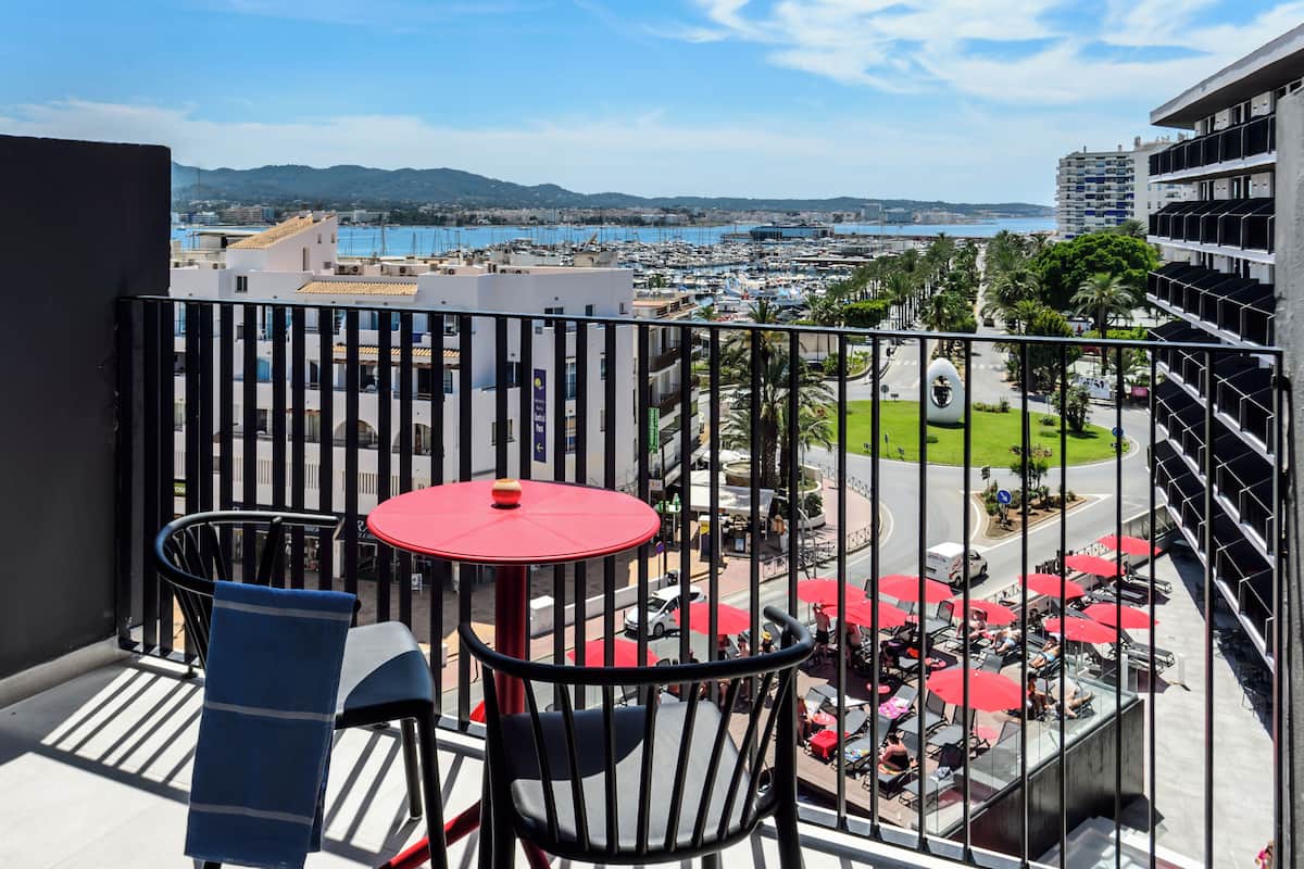 A balcony with a red table and chairs, overlooking a marina with boats and a cityscape in the distance.