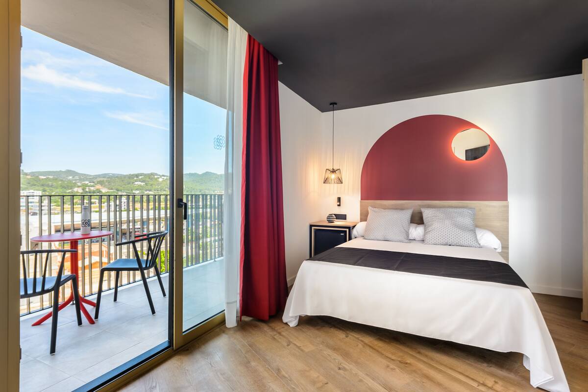 A hotel room with a bed, a nightstand, a lamp, a red curtain, and a balcony with chairs and a table.